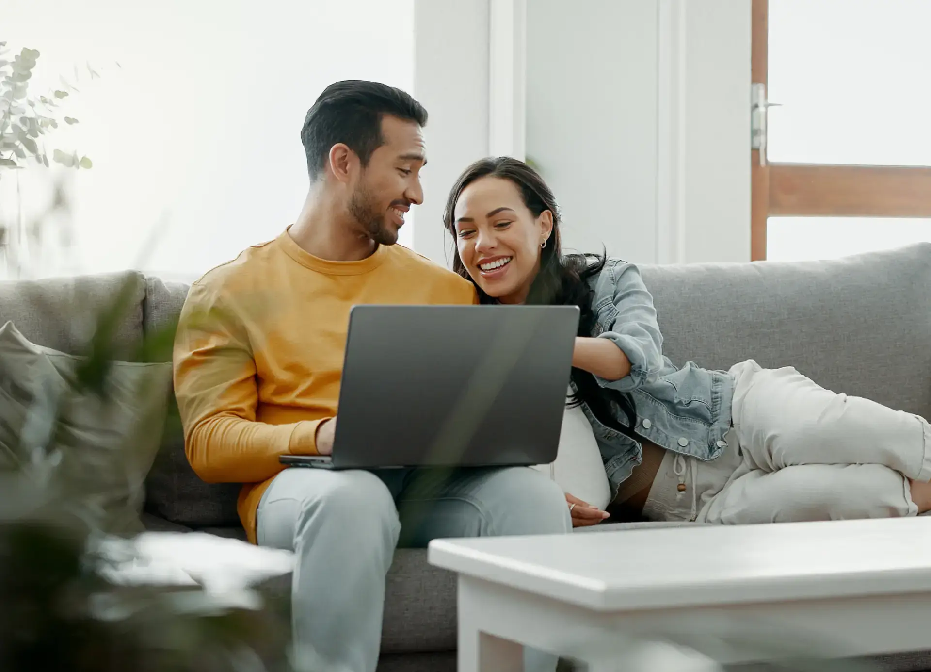 A smiling couple sits together on a gray couch looking at a laptop screen in a bright living room.